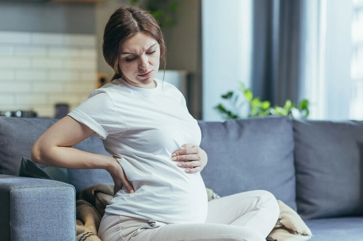 کمر درد بارداری - خانم باردار با درد کمر نشسته بر روی مبل 