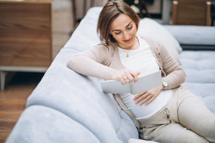 کمر درد بعد از زایمان - زن باردار در حال مطالعه کتاب نشسته بر روی مبل 